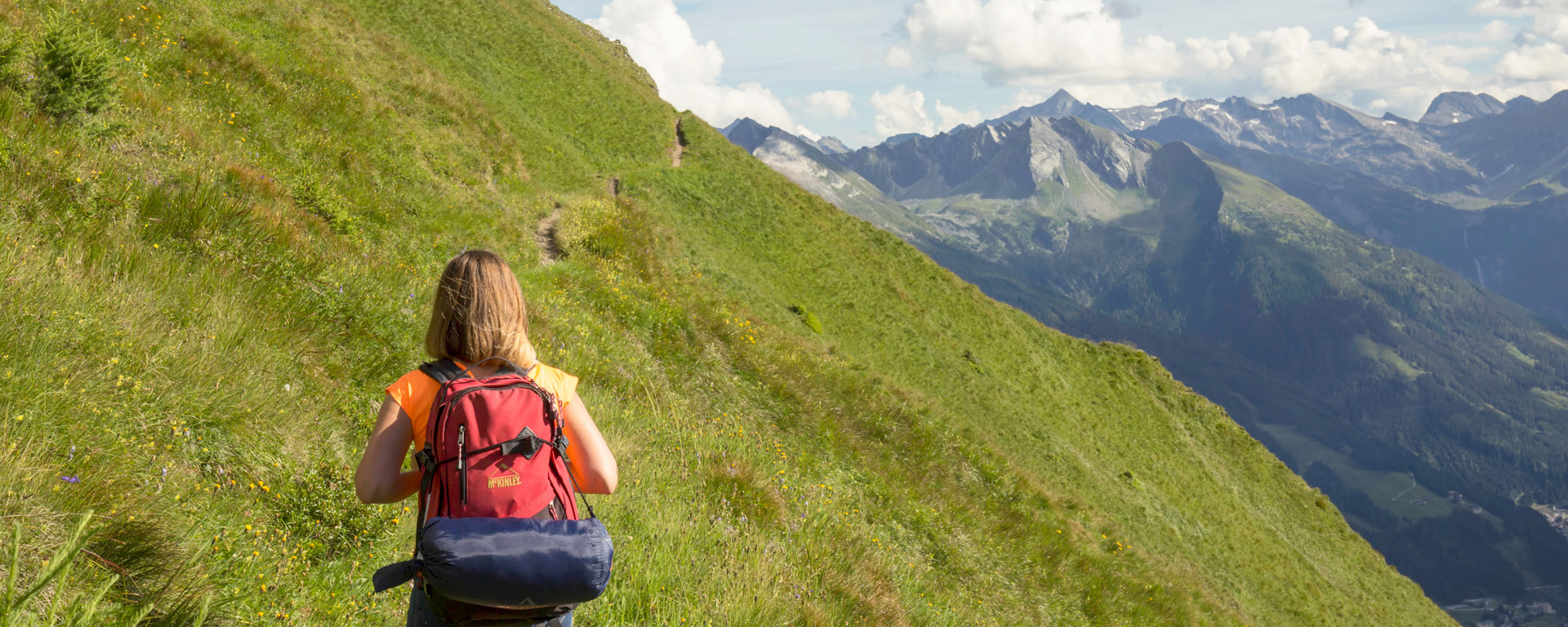 Ein Wanderer geht entlang einer grünen Alm, in der Ferne das umliegende Bergpanorama von Gastein.